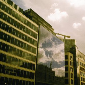 A modern office building with a glass facade reflecting clouds and surrounding structures, shot on Harman Phoenix II 200 film. The image has a warm, pinkish-yellow cast and visible grain, giving it a distinctive retro aesthetic.
