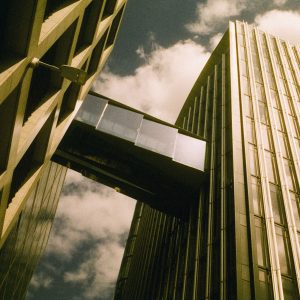 A dramatic upward view of two modern glass and steel buildings connected by a skybridge, photographed on Harman Phoenix II 200 film. The image has warm, slightly pinkish tones with noticeable grain, capturing a partly cloudy sky between the structures.