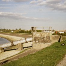 A sunny lakeside path with geese walking along the gravel track beside the water. A large metal pipe and concrete structure with graffiti sit near the shoreline, with trees and buildings visible in the distance under a sky of scattered clouds.